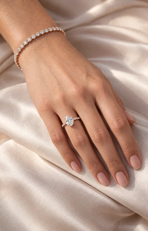 Woman wearing a rose-gold oval diamond ring and diamond tennis bracelet, with her hand resting on blush silk.