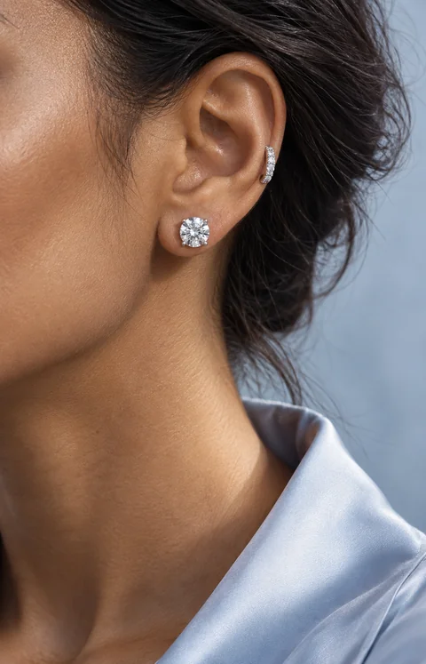 Close-up of a woman wearing a round diamond stud and a small white-gold diamond huggie, with a pale blue collar.