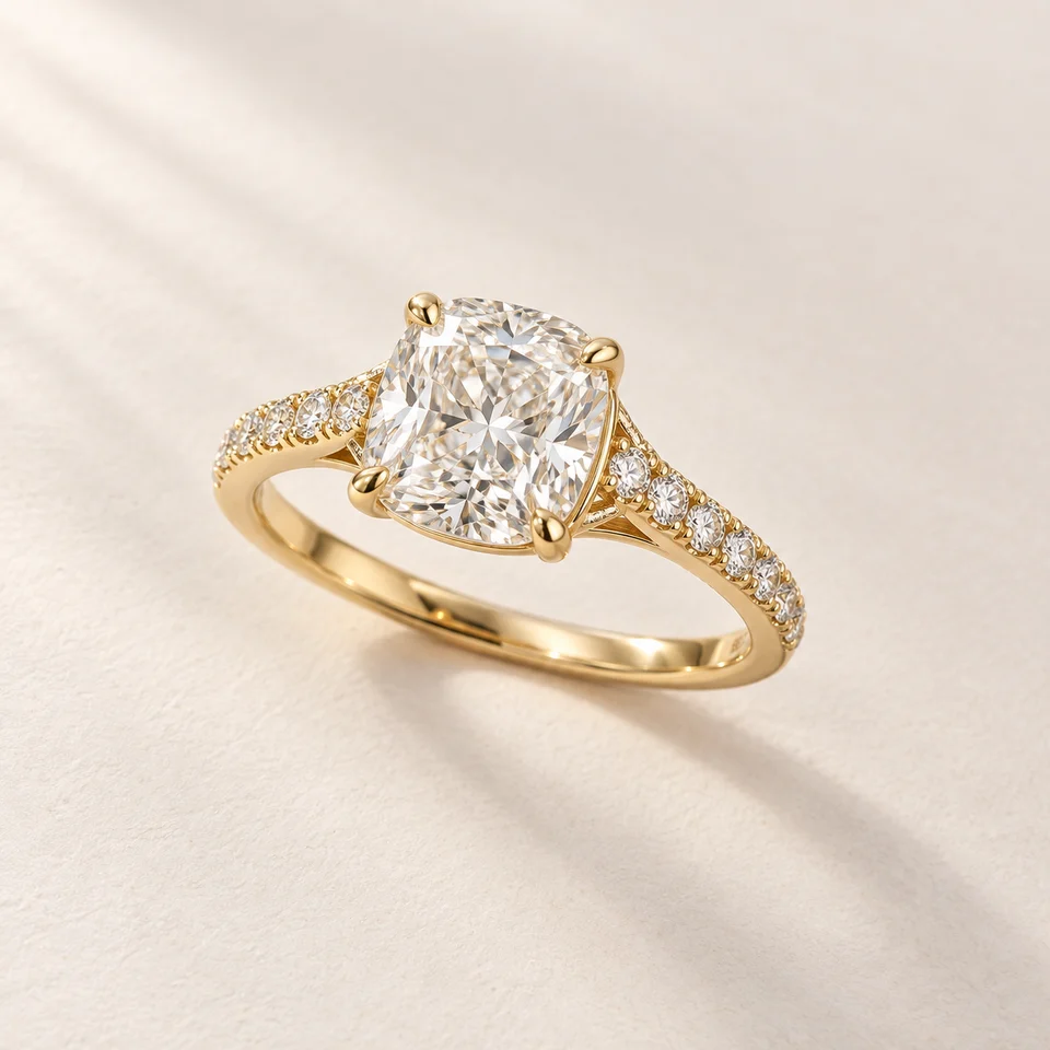 Close-up of a yellow-gold cushion-cut diamond ring with four claw prongs, diamond-pavé shoulders and a complete polished band on an ivory surface.