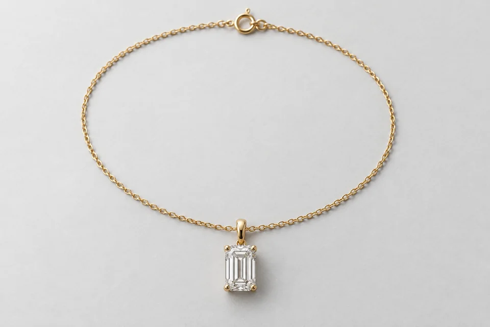 A yellow-gold emerald-cut diamond pendant with its entire delicate chain arranged in a closed oval on pale paper.