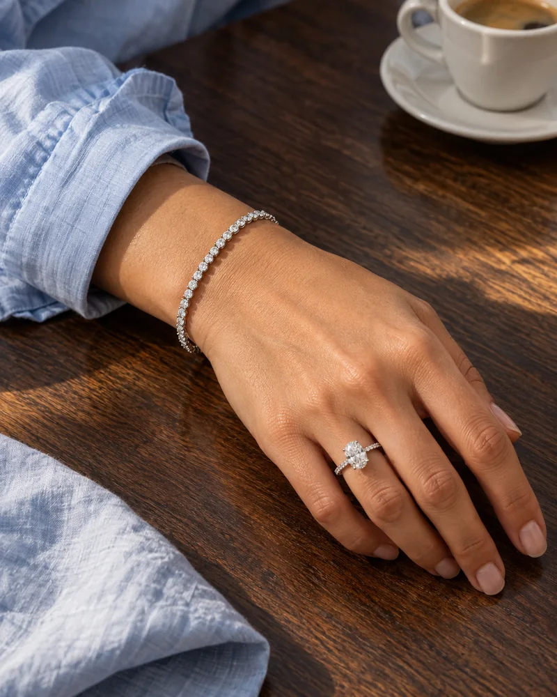 A relaxed hand wearing an oval diamond ring and white-gold diamond tennis bracelet beside a pale blue shirt cuff on a walnut café table.