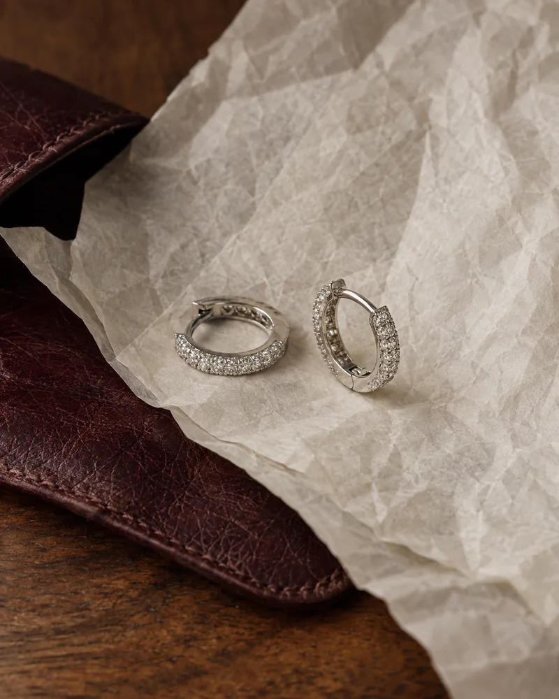 Two white-gold diamond huggie earrings resting on ivory tissue over a burgundy leather pouch.