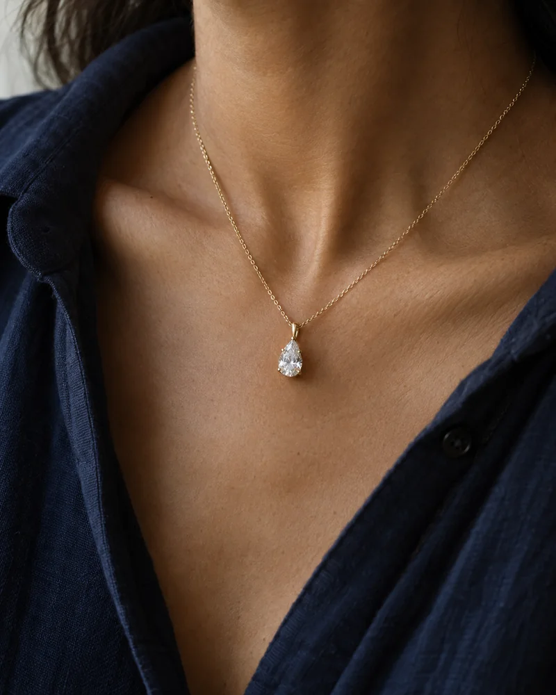 Close-up of a pear diamond pendant on a fine yellow-gold chain against an open navy cotton shirt.