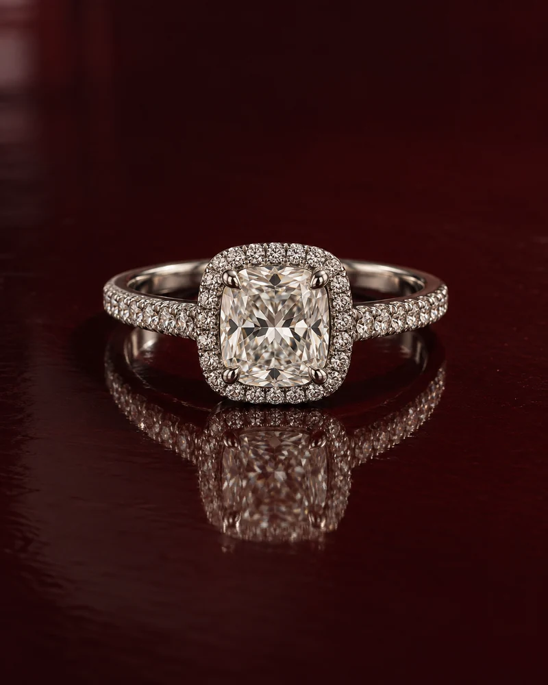 A white-gold cushion-cut diamond halo ring on dark oxblood lacquer with a soft reflection and directional evening light.