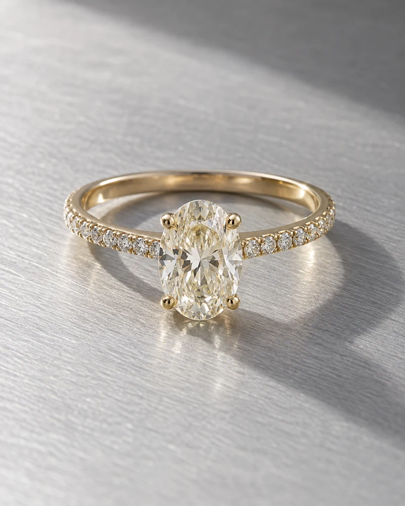 A yellow-gold oval diamond ring with pavé shoulders on cool brushed metal, lit softly from the side.