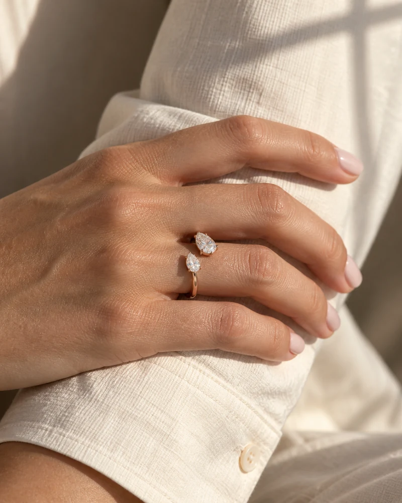 A relaxed hand wearing a rose-gold ring with two pear diamonds beside a cream cotton cuff in afternoon sunlight.