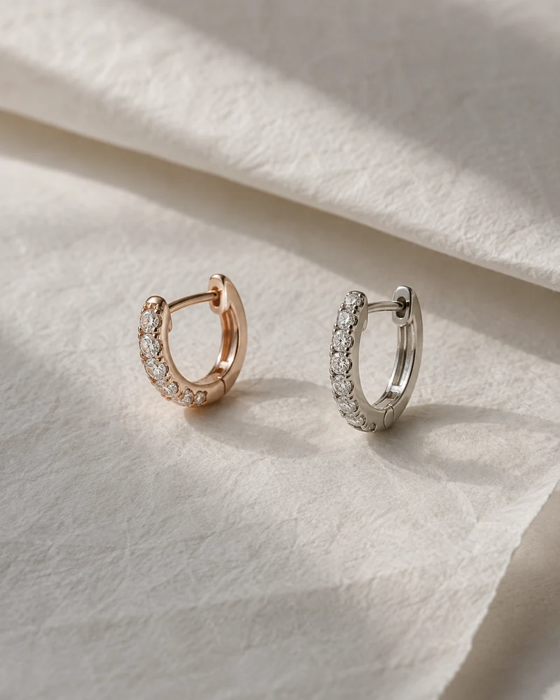 A rose-gold and a white-gold diamond huggie earring, gently separated on folded off-white tissue.