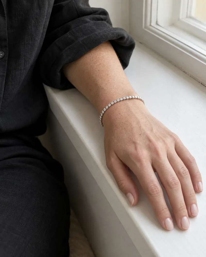 A fine white-gold diamond tennis bracelet worn beside a rolled black linen cuff, with a relaxed hand on a white windowsill.