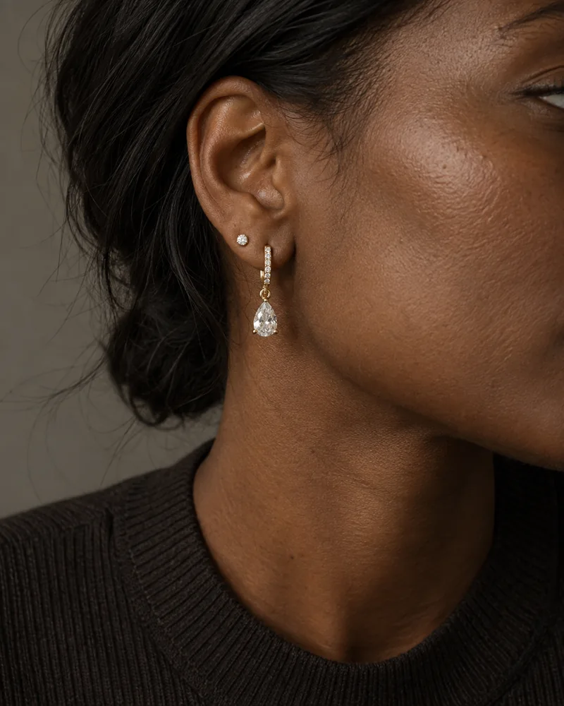 A yellow-gold diamond huggie with a pear diamond drop and a small diamond stud worn with loosely tucked-back hair.