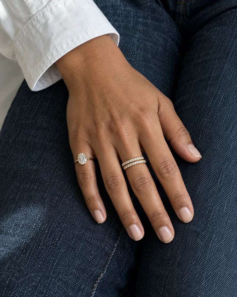 A relaxed hand wearing yellow-gold diamond bands and an oval diamond ring against dark denim and a white shirt cuff.